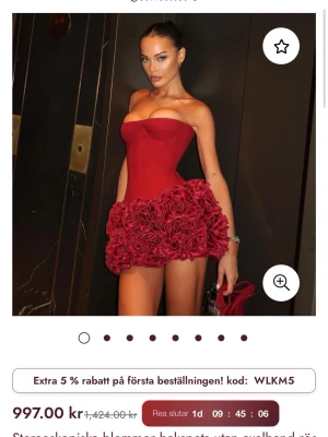 Röd axelbandslös klänning med rosvolang - Axelbandslös röd klänning med markerad bustier och volangdetaljer i form av stora rosor längs nederkanten. Klänningen har en tight passform och kort längd, perfekt för att sticka ut. Materialet ser ut att vara polyester med satinfinish.