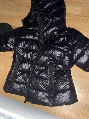 Svart glansig pufferjacka med huva - Säljer en svart, glansig pufferjacka med huva och två dragkedjeförsedda fickor framtill. Jackan har quiltad design och hög krage för extra värme. Perfekt för kalla vinterdagar och riktigt snygg till streetwear-stilen.