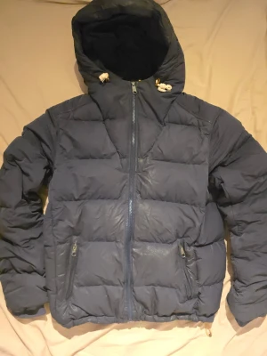 Peak Performance Frost Down Hood Limited Edition – Feather Print – M - Säljer en Peak Performance Frost Down Hood Limited Edition med ovanligt feather print och 1986 heritage patch. Marinblå med gula detaljer. Varm och skön dunjacka med huva, perfekt för höst/vinter. Bra skick utan hål eller större slitage. Storlek M.