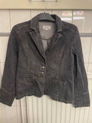 Svart jeansjacka  - Stilren svart jeansjacka från Cappuccini med klassisk krage, dekorativa knappar framtill och två halv stora fickor. Jackan har figurnära passform och detaljerade sömmar som ger en cool look. Tillverkad i bomull med snygga knappdetaljer även vid ärmsluten.