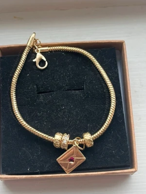 Guldigt armband med berlock och pärlor - Snyggt guldigt armband med ormkedja och dekorativa pärlor. Armbandet har en fyrkantig berlock med en röd sten i mitten och detaljerade pärlor på sidorna. Stängs med karbinhake och levereras i ask. Perfekt accessoar för att lyfta din stil.