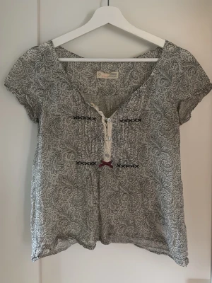 Mönstrad blus från Odd Molly - Grå/grön och vit blus från Odd Molly med paisleymönster, korta ärmar och v-ringning. Dekorativa knappar och broderidetaljer framtill. Tunn och luftig i materialet. Har en liten grön fläck på ena sidan av v-ringninhen, men syns inte så mycket (se bild).