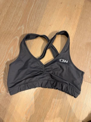 Grå sport-bh från ICANIWILL - Snygg grå sport-bh från ICANIWILL med korsade axelband i ryggen och diskret rynkning framtill. Tillverkad i stretchigt funktionsmaterial som passar perfekt för träning. Logga på bröstet ger en sportig look.