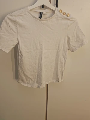 Vit t-shirt med guldknappar från Zara - Vit t-shirt från Zara med korta ärmar och tre dekorativa guldknappar på ena axeln. Klassisk rund halsringning och rak passform. Tillverkad i mjuk bomull som känns skön mot huden. Perfekt för en clean och stilren look.
