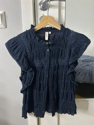 Mörkblå volangblus  - Mörkblå blus från H&M med korta volangärmar och knappar framtill. Blusen har en krinklad struktur och dekorativa spetsdetaljer längs axlar och bröst. Tillverkad i ett lätt och luftigt tyg, perfekt för en trendig och avslappnad look.