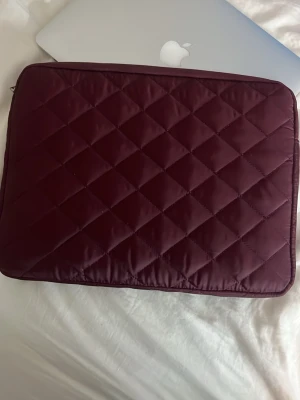 Quiltad vinröd datorväska - Snygg quiltad datorväska i vinrött satinliknande material med rutmönster. Perfekt för att skydda din laptop och ge en stilren look. Dragkedja längs kanten och mjuk vaddering för extra skydd. Inte använd har bara låtit den ligga. Kommer aldrig till användning så tänker att jag ska sälja men väldigt snyggt.