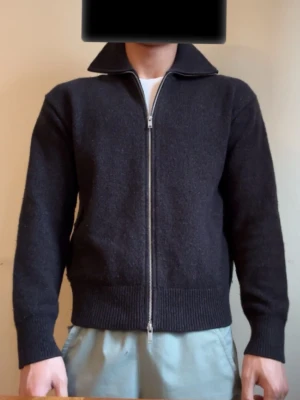 Svart ziptröja med hög krage - Säljer en svart tröja med dragkedja och hög krage. Tröjan har ribbade muddar vid ärmslut och nederkant, samt en rak passform. Perfekt för dig som gillar en stilren och enkel look. Materialet ser ut att vara ull eller en ullmix.
