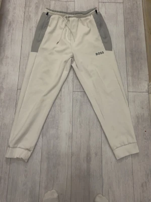 Vita mjukisbyxor från BOSS - Snygga vita mjukisbyxor från BOSS med grå detaljer vid fickorna och midjan. Byxorna har resår och dragsko i midjan samt muddar vid bensluten. Perfekta för en chill och avslappnad stil.