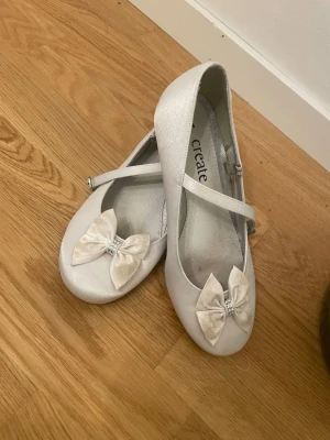 Vita ballerinaskor med rosett - Snygga vita ballerinaskor från Create med rund tå och dekorativa rosetter framtill, prydda med små glittriga stenar. Skorna har en klassisk Mary Jane-rem med spänne och låg klack. Materialet är glansigt och ger en elegant look.