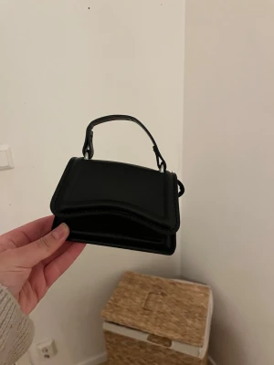 Svart minihandväska - Snygg svart minihandväska med kort handtag och stilren design. Väskan har en fyrkantig form och är gjord i läderimitation. Perfekt accessoar för att lyfta din outfit.