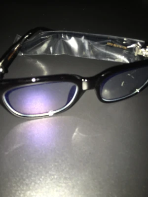 Svarta solglasögon med kristalldetaljer - Snygga svarta solglasögon från Chrome hearts med rektangulära bågar och kristalldetaljer på sidorna. Glasen har en mörk ton som ger ett coolt intryck. Perfekt för dig som vill ha en lyxig accessoar till sommaren. Fraktar inom 24 timmar