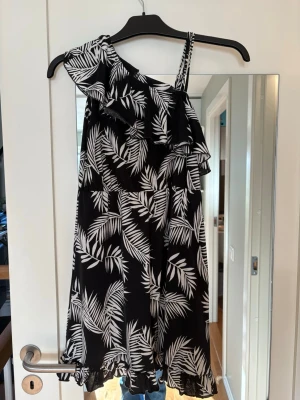 Svart one shoulder klänning med volang - Snygg svart kortklänning med vitt palmbladsmönster och volangdetaljer. Klänningen har one shoulder-design med ett smalt axelband och volang längs överdelen samt nederkanten. Perfekt för sommarens alla tillfällen. 