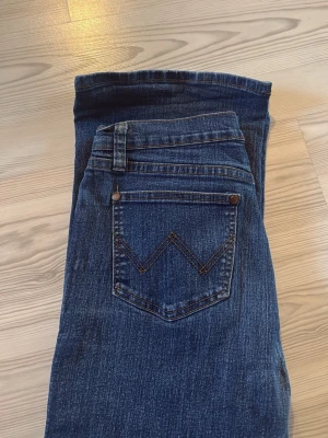 Blå bootcut jeans från Wrangler - Säljer ett par klassiska blå bootcut jeans från Wrangler med snygga kontrastsömmar och ikonisk W-detalj på bakfickan. Jeansen har normal midja, fem fickor och är tillverkade i slitstark denim. Perfekta för en avslappnad och tidlös look.