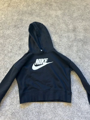 Svart cropped hoodie från Nike - Snygg svart cropped hoodie från Nike med stor vit logga på bröstet. Hoodien har huva och långa ärmar, tillverkad i mjukt bomullsmaterial. Perfekt för en sportig och avslappnad stil.