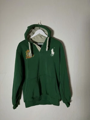 Hoodie från Polo Ralph Lauren - Snygg grön hoodie från Polo Ralph Lauren med vit broderad polo logga på bröstet och ett färgglatt märke på vänster sida och en broderad 3 på höger axel . Hoodien har en stor ficka framtill, ribbade muddar och tjocka vita dragsnören i huvan. Insidan av huvan är ljusgrå. Hoodien är ond school men  har inga defekter och inte synligt använd. Skriv gärna vid frågor så ska jag se till att svara snabbt ☺️