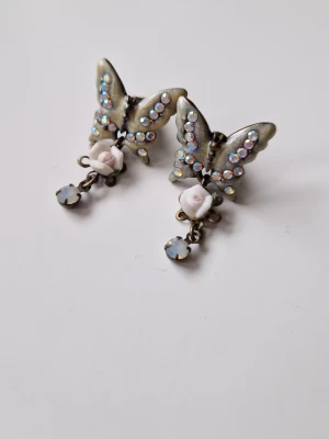 Vintage örhängen med fjärilar och strass - Unika örhängen i metall med fjärilsform, dekorerade med skimrande strass och små vita rosor i porslin. Hängande detalj med ljus sten ger extra bling. Perfekt för dig som gillar romantisk och retro stil. De är 2.5 cm långa. Oanvända och inköpta i Paris tidigt 2000-tal 