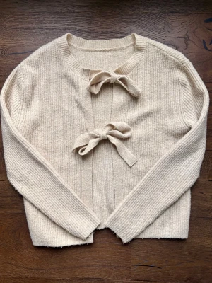 Kofta - Söt beige kofta i ribbstickat material med två dekorativa rosetter framtill som knäppning. Koftan har rund halsringning och långa ärmar. Perfekt för en trendig och mysig look.