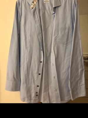 Ljusblå skjorta från Oscar Jacobson - Klassisk ljusblå skjorta från Oscar Jacobson med diskret randigt mönster. Skjortan har lång ärm, bröstficka och knappar framtill. Regular fit och tillverkad i ett skönt, strykfritt bomullsmaterial. Perfekt för en clean och stilren look.