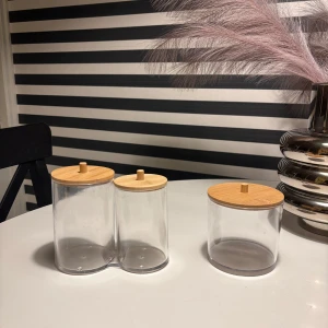Två genomskinliga burkar med trälock - Säljer två genomskinliga förvaringsburkar i plast med ljusa trälock. En burk är dubbel och sitter ihop, den andra är enkel. Perfekta för småsaker eller snacks. Snygg och stilren design som passar i de flesta hem.