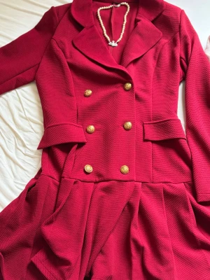 Röd kavajklänning med guldknappar - Stilren röd kavajklänning med dubbelknäppning i guld och markerad midja. Klänningen har långa ärmar, breda slag och dekorativa fickor framtill. Kjoldelen är veckad för extra volym och materialet har en strukturerad yta.