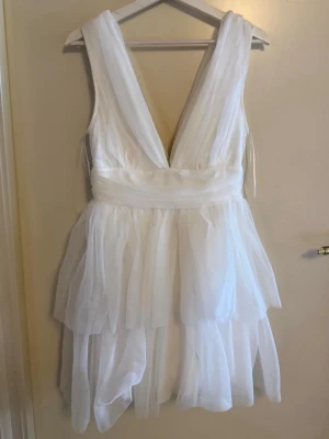 Studentklänning - Snygg vit kortklänning med omlott-design och flera lager volanger. Djup v-ringning och bred midjedetalj ger en feminin siluett. Klänningen är ärmlös och tillverkad i ett tunt, skirt material som ger ett luftigt intryck. (Aldrig använt)