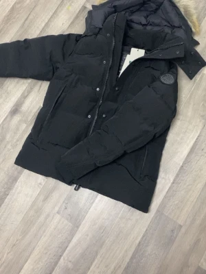 Svart pufferjacka från Canada Goose - Säljer en svart pufferjacka från Canada Goose med avtagbar huva och pälsdetalj. Jackan har ett klassiskt quiltat mönster, logopatch på ärmen och dragkedja med knappar. Perfekt för kalla vinterdagar och riktigt varm.