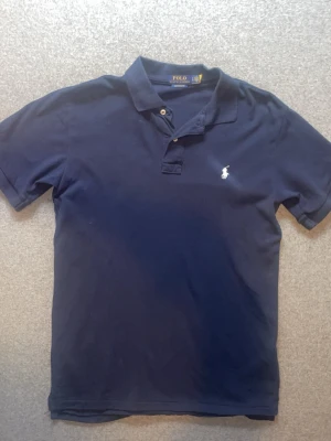 Marinblå pikétröja från Ralph Lauren - Klassisk marinblå pikétröja från Ralph Lauren med vit broderad logga på bröstet. Tröjan har korta ärmar, krage och knappar vid halsen. Perfekt för en clean och stilren look.