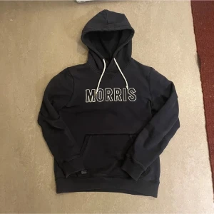 Marinblå hoodie från Morris med tryck - Snygg marinblå hoodie från Morris med vit broderad logga på bröstet och vita dragsnören i huvan. Klassisk känguruficka framtill och ribbade muddar vid ärmslut och nederkant. Perfekt för en avslappnad och trendig stil.