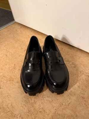 Chunky svarta loafers från Bianco - Säljer ett par chunky svarta loafers från Bianco i glansigt skinn. Skorna har en klassisk penny loafer-design med bred sula och markerad klack. Perfekta för dig som vill ha en edgy och stilren look till vardagsoutfiten.