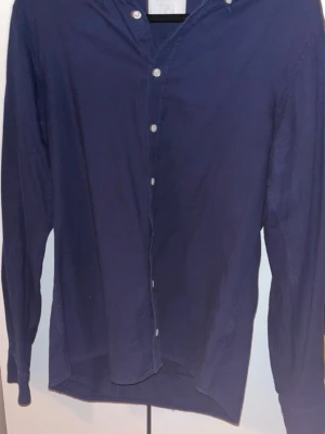 Mörkblå skjorta från Oscar Jacobson - Stilren mörkblå skjorta från Oscar Jacobson med klassisk krage och vita knappar. Skjortan har lång ärm och är tillverkad i ett mjukt bomullstyg. Perfekt för dig som gillar en clean och tidlös look.