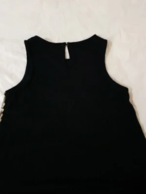 Svart ärmlös topp från KappAhl - Snygg svart ärmlös topp från KappAhl med rund halsringning och knappdetalj i nacken. Perfekt för att styla med olika outfits. Tillverkad i mjukt tyg som känns skönt mot huden.