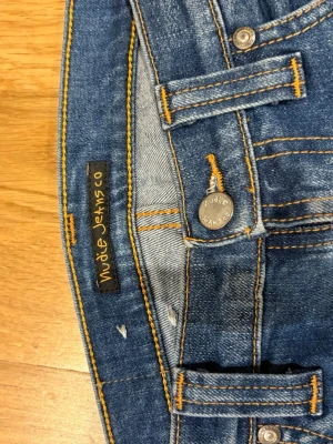 Blå smala jeans från Nudie Jeans - Säljer ett par klassiska blå jeans från Nudie Jeans med smal passform och snygga slitningar. Jeansen har orange kontrastsömmar, knappgylf och fem fickor. Perfekta för dig som gillar en tidlös och avslappnad stil. Vintage nudie jeans 😎priset inte hugget i sten !!!