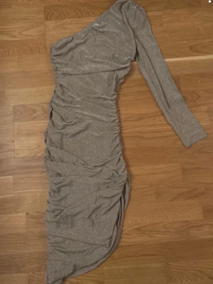 Glittrig oneshoulder långklänning - Säljer en glittrig långklänning i silver med oneshoulder-design och lång ärm. Klänningen har draperade detaljer längs sidorna som ger en snygg effekt. Perfekt för dig som vill sticka ut med en unik och trendig look.