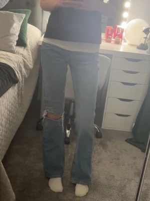 Blå bootcut jeans med slitning - Säljer ett par blå bootcut jeans med slitning vid knät. Jeansen har hög midja och klassisk femficksdesign. Perfekta för en avslappnad och trendig look. Materialet är denim i bomull. Passar dig som gillar en lite vidare passform nedtill.