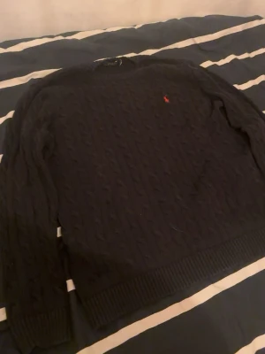 Svart kabelstickad Polo Ralph Lauren - Svart kabelstickad tröja från Polo Ralph Lauren med klassisk rund hals och ribbade muddar. Tröjan har ett rött broderat Polo-logo på bröstet och är gjord i mjukt stickat material. Perfekt för dig som gillar stilrena och tidlösa plagg.