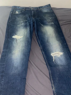 Blå slitna jeans från Hoodrich  - Säljer ett par blå jeans från hoodrich med slitningar och trasiga detaljer på benen. Jeansen har en klassisk femficksdesign och tydliga blekta partier. Perfekt för dig som gillar en cool och avslappnad look.