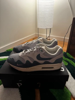 Nike x Patta Air Max 1 grå/vit - Nike x Patta Air Max 1 sneakers i grått och vitt med vågig design på ovandelen. Skorna har mesh och mockadetaljer, synlig Air Max-sula och Patta-logga på tungan. Perfekta för dig som gillar streetwear och unika samarbeten.