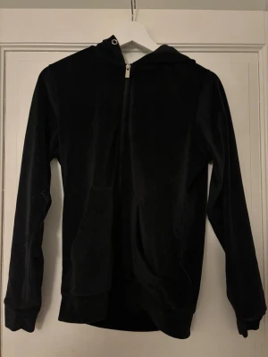 Svart velourhoodie med dragkedja - Snöret till luvan är tyvärr borta. Snygg svart hoodie i mjuk velour med dragkedja framtill. Klassisk passform med långa ärmar och ribbade muddar. Perfekt för en chill och avslappnad stil. Huvan ger extra cozy känsla.