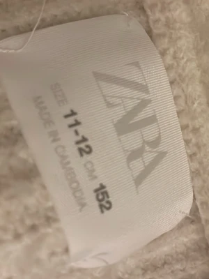 Stickad beige/vit kofta från Zara - Mysig vit kofta från Zara med stora bruna knappar framtill. Koftan har en djup V-ringning och är stickad i ett mjukt material. Perfekt att slänga över en topp för en chill look.