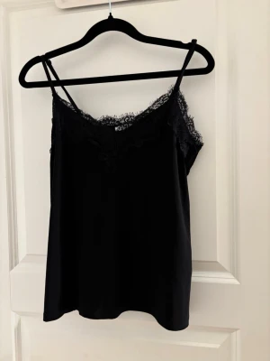 Svart linnetopp med spetskant - Snygg svart linnetopp med tunna axelband och dekorativ spetskant längs urringningen. Toppen har en lös passform och är perfekt för att styla lager på lager. Materialet känns mjukt och följsamt mot huden.