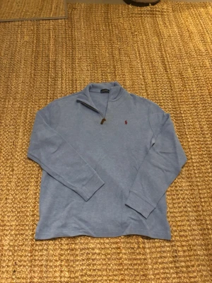 Ljusblå quarterzip från Polo Ralph Lauren - Ljusblå långärmad tröja från Polo Ralph Lauren med halvzip och liten röd logga broderad på bröstet. Tröjan har ribbade muddar vid ärmslut och är tillverkad i mjukt bomullsmaterial. Perfekt för en avslappnad men ändå stilren look. Den har en fläck på bröstet men inget som syns supermycket