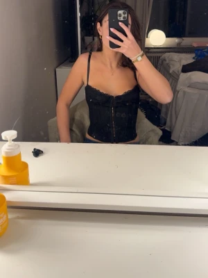 Svart spetskorsett topp - Säljer en svart korsett med tunna axelband och dekorativ spets längs överkanten. Korsetten har knäppning framtill och en figurnära passform som framhäver midjan. Perfekt för dig som vill ha en trendig och stilren look.