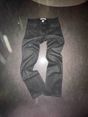 J Lindeberg waxed demin - Säljer ett par svarta jeans med rak passform och klassisk femficksdesign. Jeansen har bälteshällor och knappgylf. Materialet ser ut att vara bomull med lite stretch för extra komfort. Perfekta till en avslappnad och stilren look. Archivee Längd 36 Waist 31