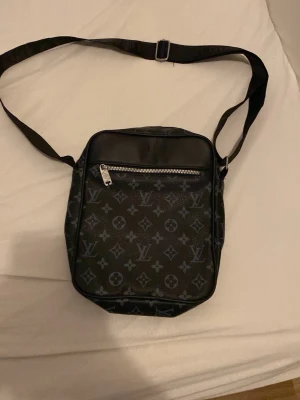 Louis Vuitton svart axelväska monogram - Snygg svart axelväska från Louis Vuitton med klassiskt monogrammönster i grått. Väskan har justerbar axelrem, en dragkedjeficka framtill och är tillverkad i tåligt canvasmaterial med detaljer i läder. Perfekt för dig som vill ha en stilren och trendig accessoar.