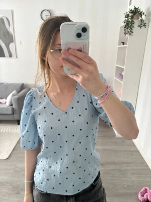 Blå prickig blus med puffärm - Söt ljusblå blus med svarta prickar och v-ringning. Blusen har puffiga korta ärmar och är tillverkad i ett lätt, strukturerat tyg som ger en trendig look. Perfekt för dig som gillar en lekfull och modern stil.