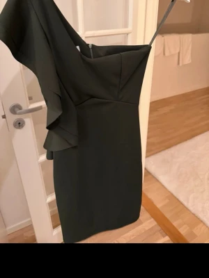 Mörkgrön one shoulder klänning - Snygg mörkgrön klänning med one shoulder-design och volangdetalj på axeln. Kort modell som sitter figurnära och har en elegant, stilren look. Perfekt för dig som vill sticka ut med en unik axelbandslös stil.