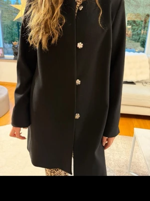 Svart klassisk kappa med blommiga knappar - Stilren svart kappa med raka linjer och dekorativa blommiga knappar framtill. Kappan har en enkel, tidlös design och är knälång. Perfekt för dig som gillar minimalistisk stil med en twist. Materialet ser ut att vara ullblandning.