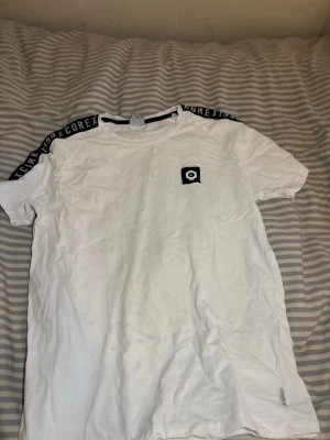Vit t-shirt från Jack & Jones Core - Snygg vit t-shirt från Jack & Jones Core med svart logga på bröstet och svarta band med text på axlarna. Klassisk rund hals och korta ärmar. Perfekt till jeans eller shorts för en clean och sportig look.