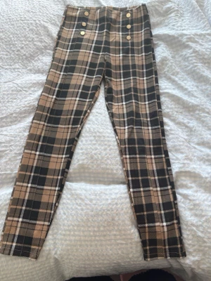Rutig byxa med knappar från Zara - Snygga rutiga byxor från Zara i beige, svart och vitt med dekorativa guldknappar byxorna är perfekta för barn som gillar trendiga mönster och detaljer. Tillverkade i mjukt material för en bekväm känsla.