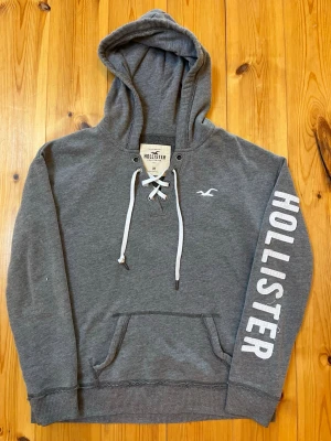Grå hoodie från Hollister med tryck - Snygg grå hoodie från Hollister med vit snörning i halsen och stor ficka framtill. På ena ärmen står det HOLLISTER i vitt tryck och loggan finns broderad på bröstet. Tillverkad i mjuk bomullsblandning och har en klassisk huva. 